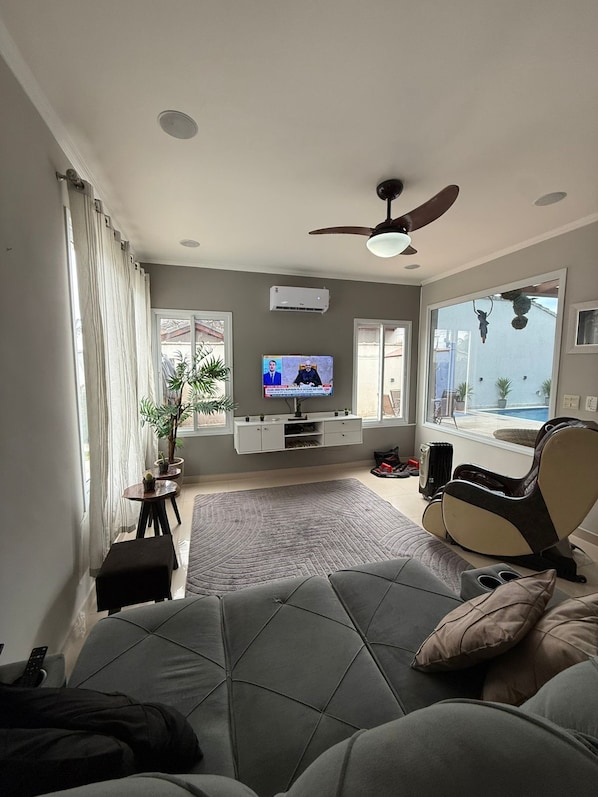 A modern living room with a sofa, a TV, a ceiling fan, and a view of the outdoors. - Beautiful two-story house in the Morada da Praia condominium. (Bertioga)