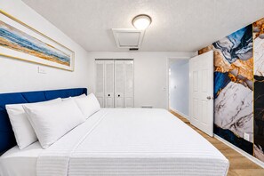 A bedroom with a bed, a blue headboard, white bedding, a ceiling light, a door with a marble-patterned wall, and a hallway. - Private Pool Retreat in Tampa | 5BR/5BA Vacation Home (Tampa)