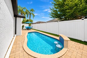 Une piscine rectangulaire dont le bord est recouvert de carreaux, entourée d’une terrasse en briques et d’une clôture blanche.