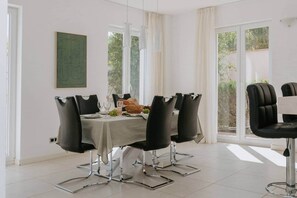 A dining area with a table set for four, black chairs, a green abstract painting, and large windows with curtains. - Villa Oleander: Pool & Rooftop Cinema (Splitska)