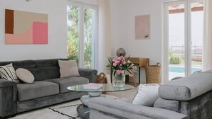 A modern living room with a grey sofa, a glass coffee table, and a view of a swimming pool. - Villa Oleander: Pool & Rooftop Cinema (Splitska)