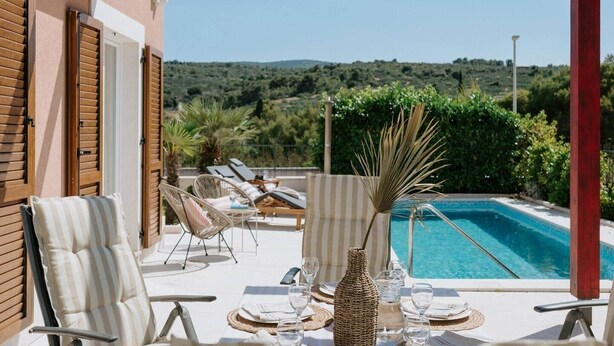 A poolside area with lounge chairs, a dining table set for two, and a view of a pool and hills in the background. - Villa Melograno: Pool & Rooftop Cinema (Splitska)