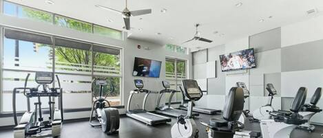 A gym with treadmills, elliptical machines, and stationary bikes, large windows, and a TV on the wall.