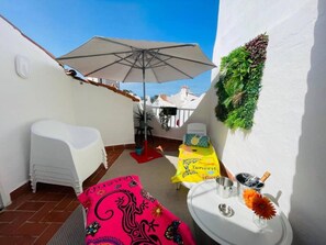 Ein kleiner Balkon mit weißem Stuhl, rotem Hocker, gelbem Handtuch, Tisch mit Flasche und Gläsern sowie einer grünen Wanddekoration.
