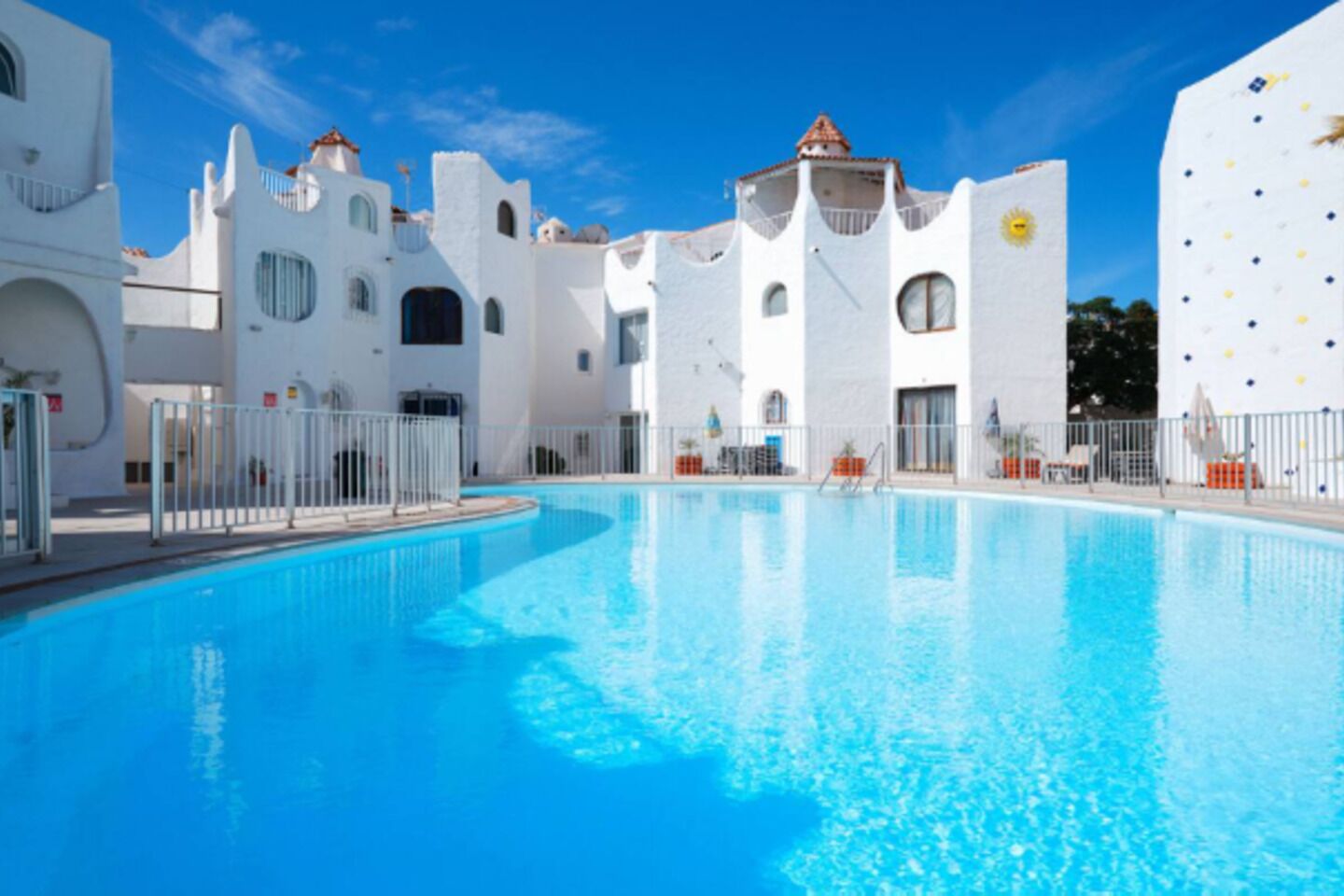 A swimming pool surrounded by white buildings with arched windows and balconies, under a clear blue sky.