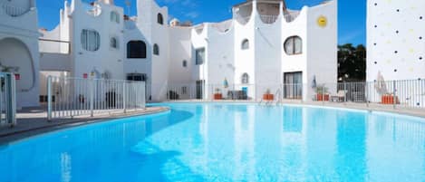 A swimming pool surrounded by white buildings with arched windows and balconies, under a clear blue sky.
