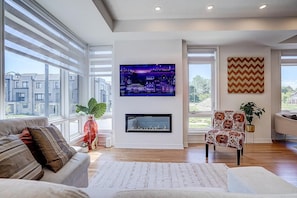 A modern living room with a fireplace, large windows, a television, and a patterned armchair.
