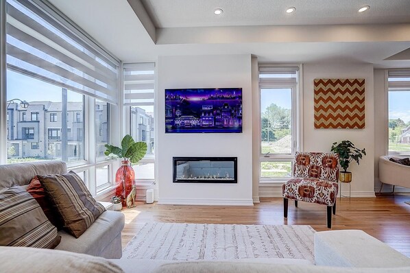 A modern living room with a fireplace, large windows, a television, and a patterned armchair.