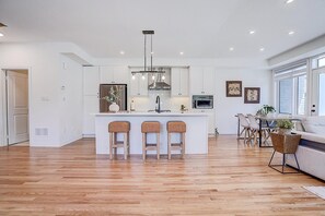 A modern kitchen with white cabinetry, a central island with stools, a dining area with a table and chairs, and a large window providing natural light.