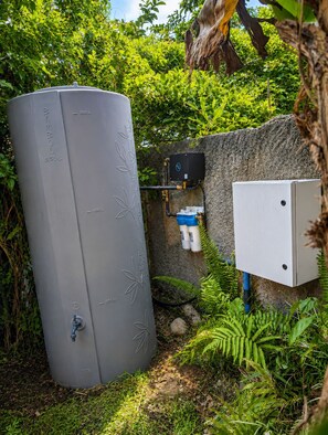 Un réservoir d’eau grise équipé d’une vanne, relié à une boîte noire et à une boîte électrique blanche, le tout installé contre un mur de pierre agrémenté de verdure.