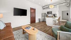 A modern living room with a flat-screen TV, a wooden coffee table, a brown leather sofa, a green armchair, and a kitchen area with a microwave and sink. - 1 BR King Suite - Freshwater Inn - Bridgman, MI (Bridgman)