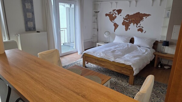 A bedroom with a bed, a desk, a chair, a world map on the wall, and a view of the outside.