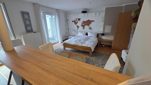 A hotel room with a wooden desk, a bed with white bedding, a world map on the wall, a small bedside table, and a wardrobe. - Cozy vacation apartment in HD-Wieblingen (Heidelberg)