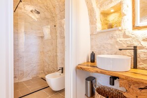Un bagno con parete in pietra, lavandino bianco, mobile contenitore in legno e bidet.