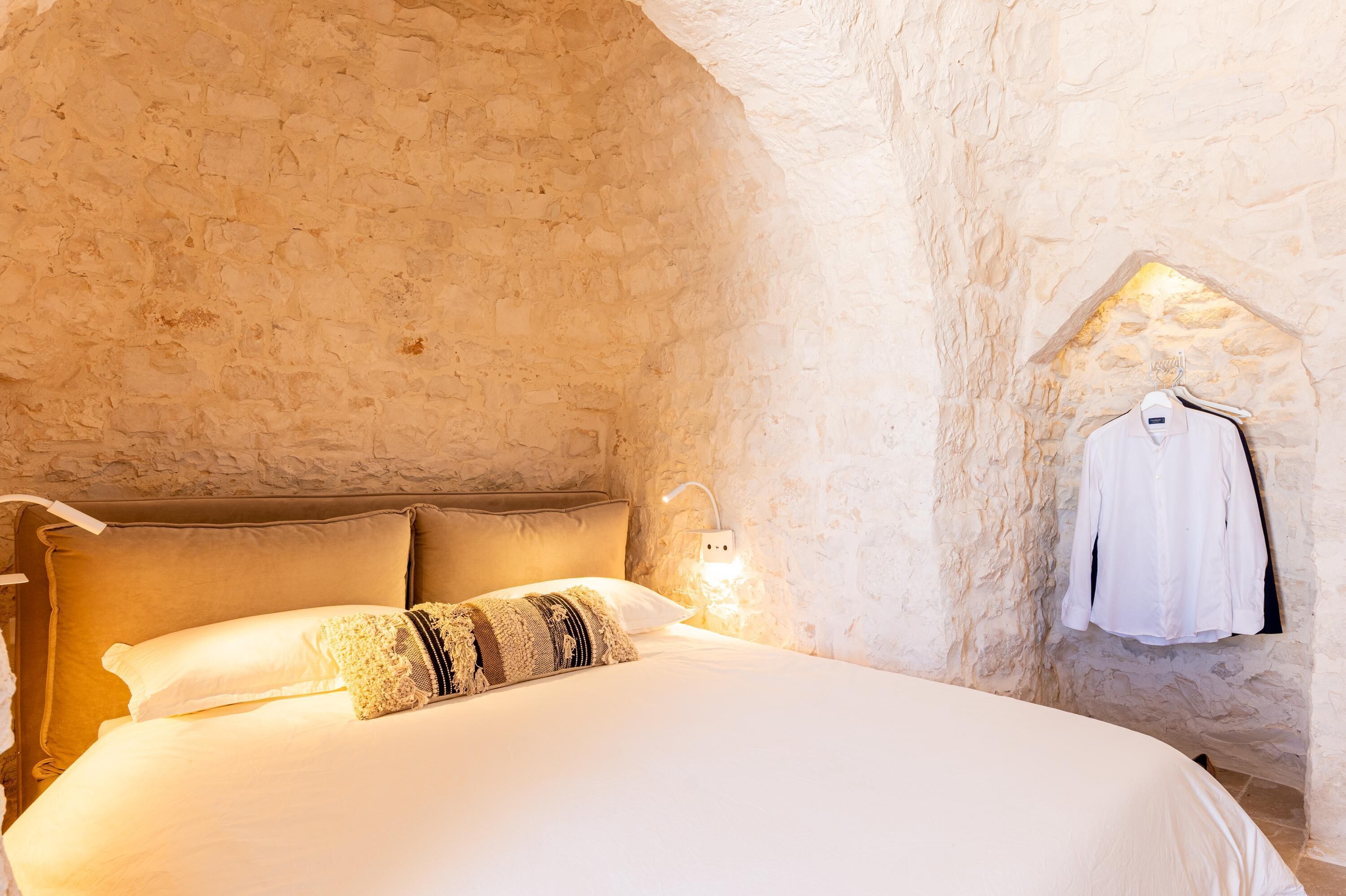 Ein Schlafzimmer mit Steinswand, einem Bett mit weißer Bettwäsche, einer Hängeleuchte und einem weißen Hemd an der Wand.