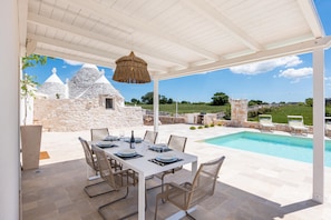 Zona pranzo coperta all'aperto con piscina e edifici in pietra sullo sfondo.