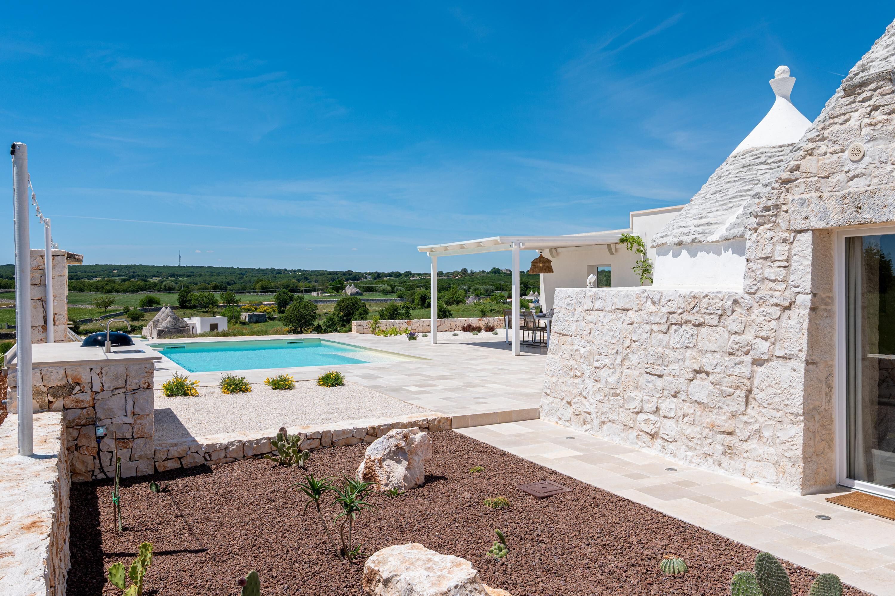 Una piscina con muro in pietra e una pergola, circondata da un giardino con rocce e piante.