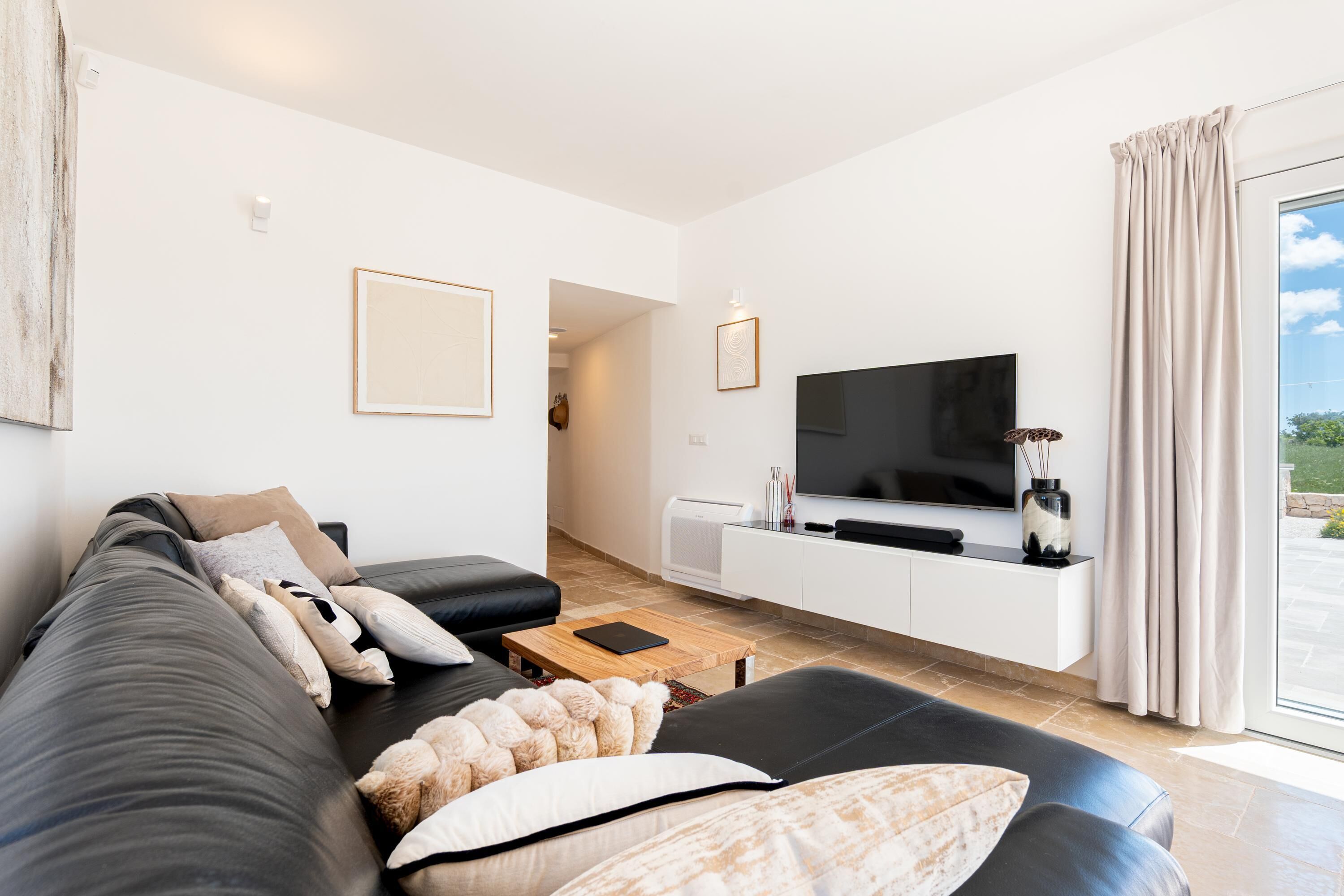 En moderne stue med en sort sofa, et fladskærms-tv, et hvidt tv-møbel og et stort vindue med gardiner.