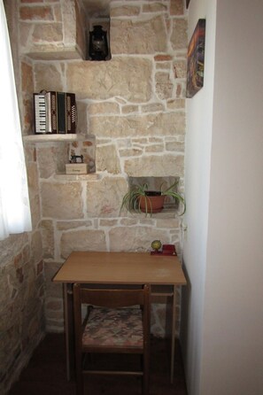 A corner room with a stone wall, a wooden table, a chair, a shelf with books, a clock, and a potted plant. - House Batel by Interhome (Barbariga)