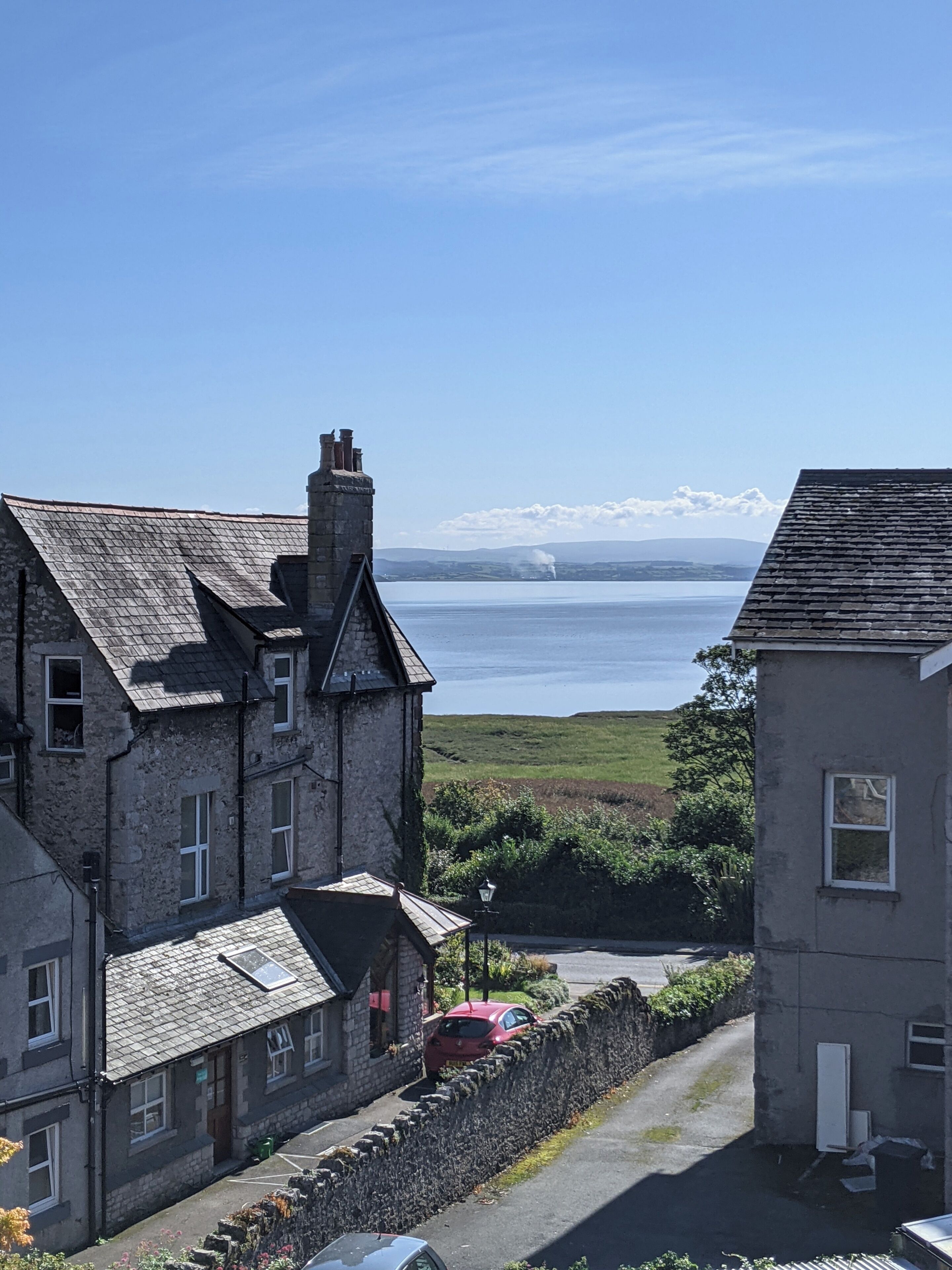 A stone building with a chimney, a red car parked in front, and a view of a body of water with mountains in the distance.