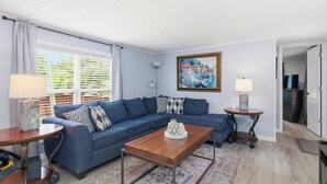 A living room with a sectional sofa, a wooden coffee table, a painting on the wall, and a view through the window.