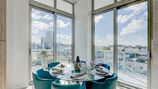 A modern dining area with a glass table, blue chairs, and a city view through large windows. - AEA - Biscayne BLVD Miami (Miami)