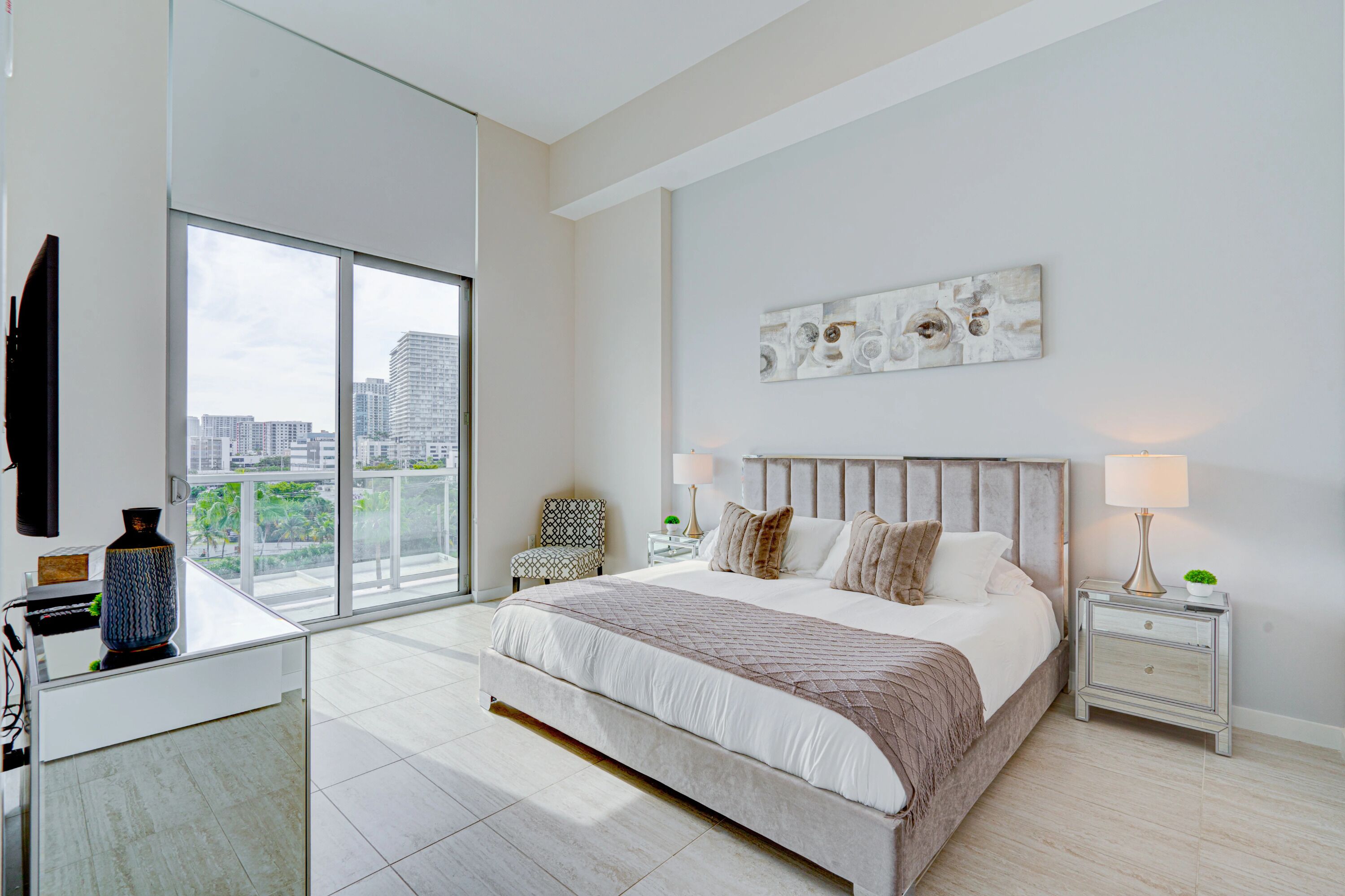 Ein modernes Schlafzimmer mit großem Bett, Blick auf die Stadt, Fernseher und einer dekorativen Vase.