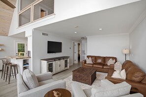 Amplia sala de estar con techo alto, comedor con mesa de madera, sofá de cuero y televisor montado en la pared.