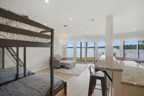 Een moderne slaapkamer met stapelbedden, een grijze bank, een keuken met een marmeren aanrechtblad en grote ramen met uitzicht op een waterpartij.