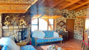 Un interior rústic amb una llar de pedra, bigues de fusta, un sofà amb dibuixos i una taula de dinar amb un cistell de pa.