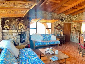 Un interior rústico con chimenea de piedra, vigas de madera, un sofá estampado y una mesa de comedor con una cesta de pan.