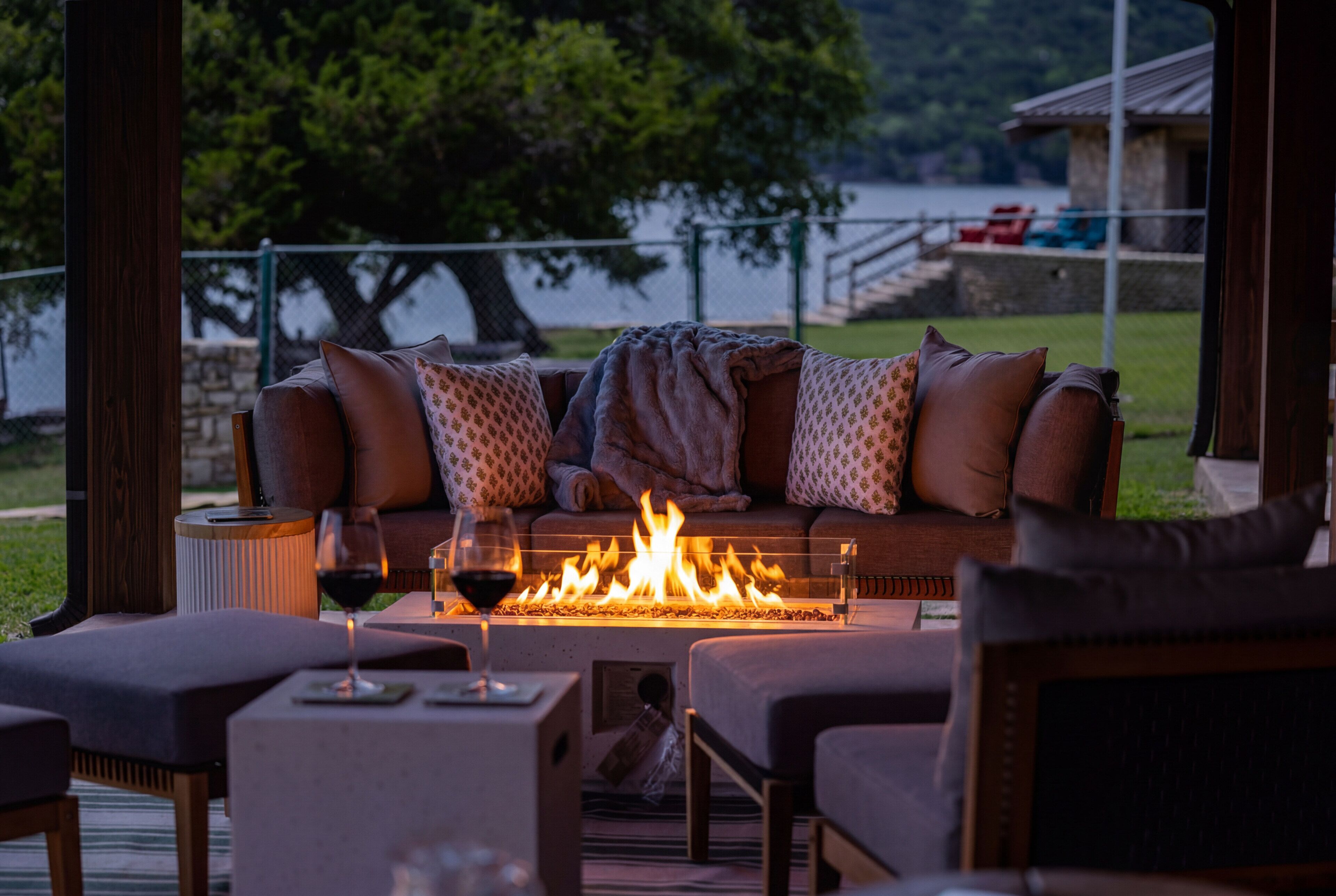 A cozy outdoor seating area with a fire feature, a glass of wine, and a view of a lake.