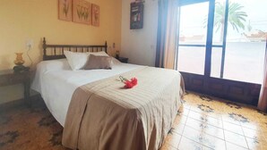 Ein Hotelzimmer mit Bett, Deckenventilator, Fenster mit Blick auf Palmen und einer Wand mit floralen Bildern.