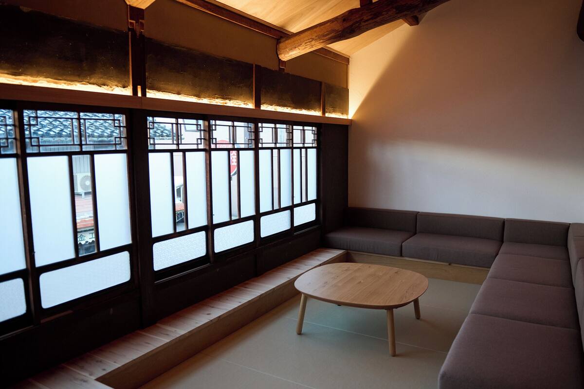 A traditional Japanese room with tatami flooring, a low wooden table, a cushioned seating area, and large windows with a view of the outside.