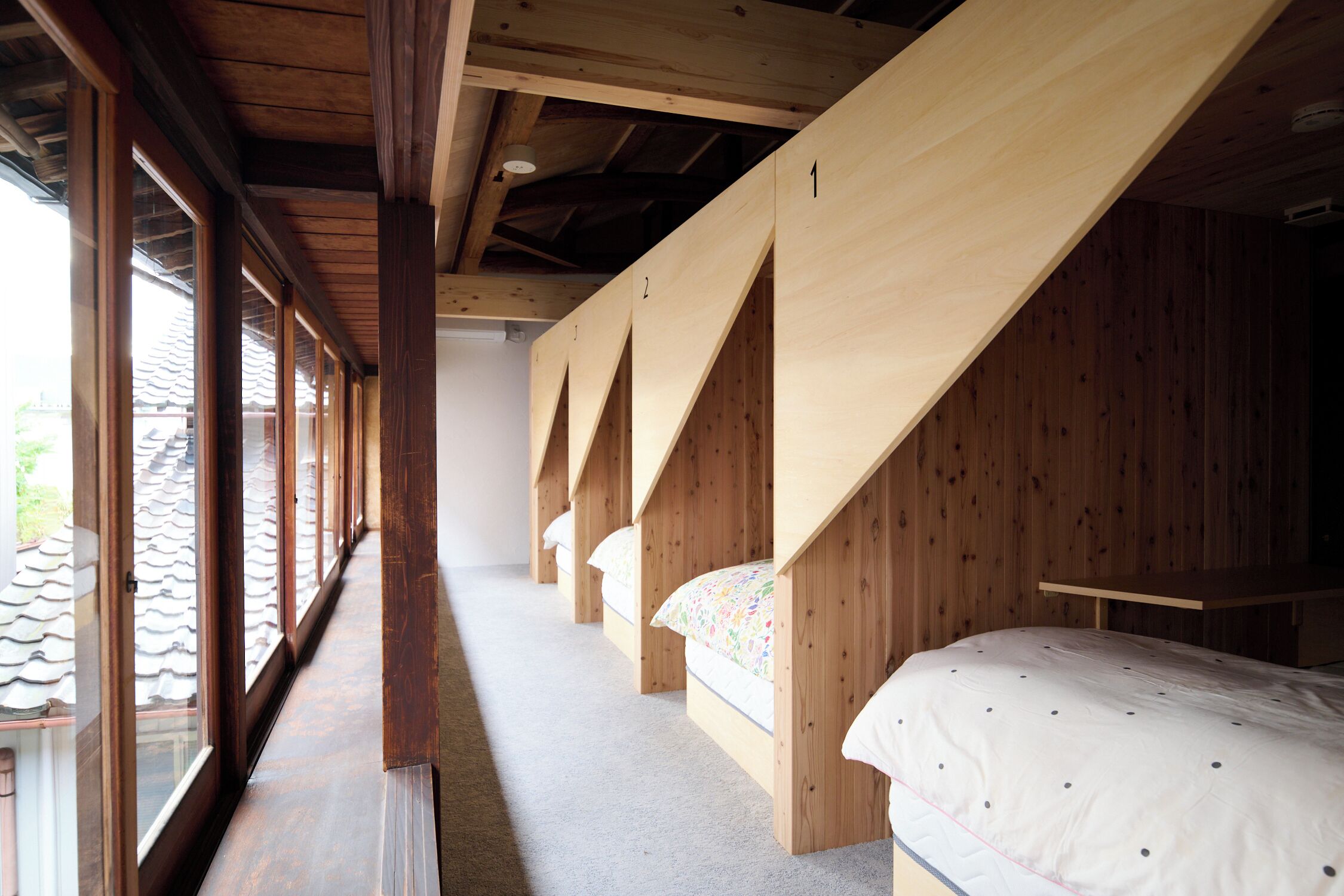 A row of wooden bunk beds with a sloped ceiling, large windows on one side, and a view of a tiled roof outside.