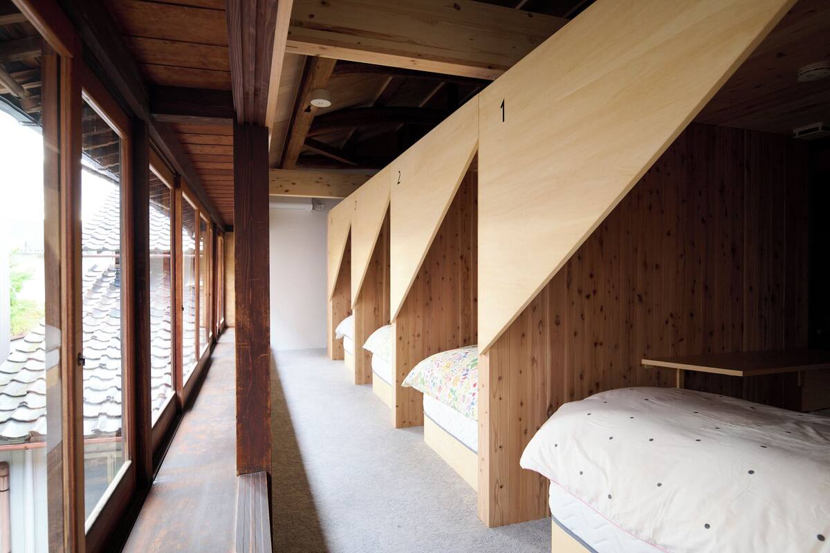 A row of wooden bunk beds with a sloped ceiling, large windows on one side, and a view of a tiled roof outside.