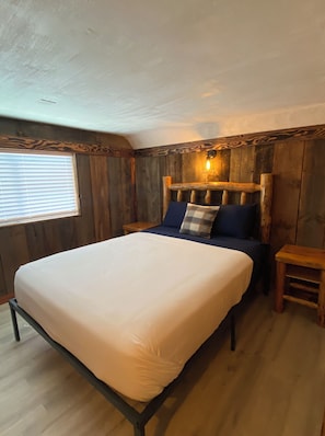 A bedroom with a large bed, wooden walls, a window, and a nightstand. - Lazy Lake Outpost (Kettle Falls)