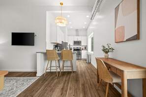 Una cocina moderna con armarios blancos, una mesa de comedor de madera, dos taburetes de mimbre y una lámpara colgante. Hay un televisor de pantalla plana montado en la pared.