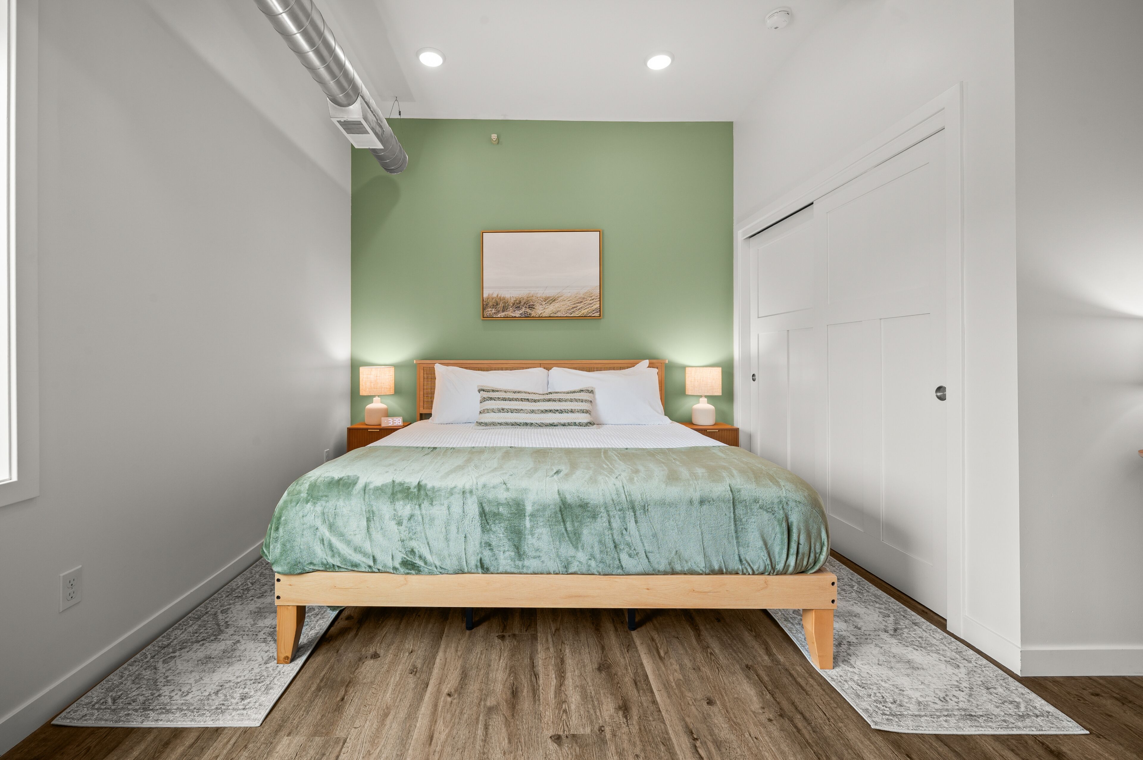 A bedroom with a bed, two bedside lamps, a green wall, a framed picture, and a white door.