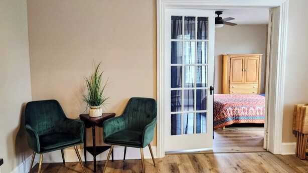 A room with two green chairs, a small table with a plant, a wooden cabinet, and a bed with a colorful bedspread.
