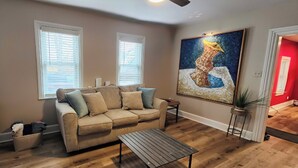 A living room with a beige sofa, two windows with blinds, a painting on the wall, and a wooden floor.