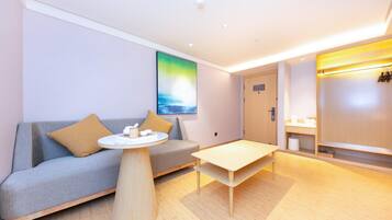 A modern hotel room with a grey sofa, a round coffee table, a wooden floor, a painting on the wall, and a wooden wardrobe.