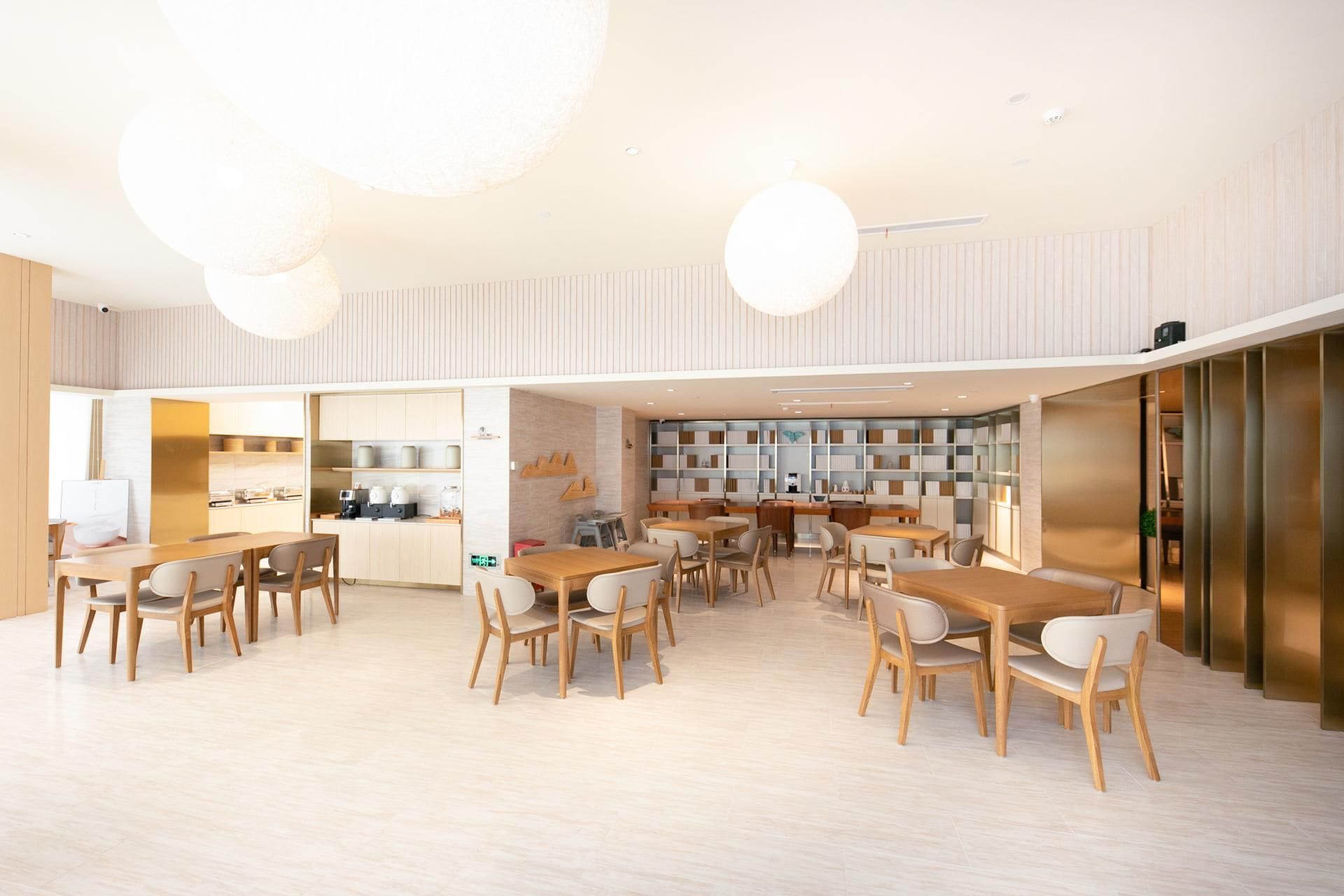 A modern interior with multiple dining tables and chairs, a kitchen area, and a bookshelf.