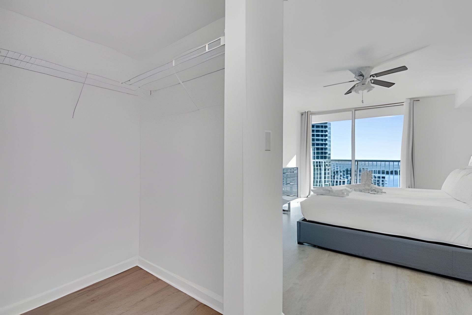 Une chambre moderne avec un grand lit, un ventilateur de plafond et une vue sur le paysage urbain, visible à travers de grandes fenêtres.