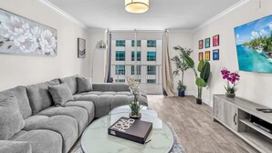 Ett modernt vardagsrum med en grå soffa, ett glasbord med en bok, ett TV-bord och ett stort fönster med gardiner.