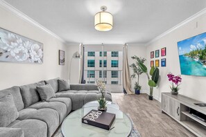 A modern living room with a grey sofa, a glass coffee table with a book, a TV stand, and a large window with curtains. - The Club (Miami)