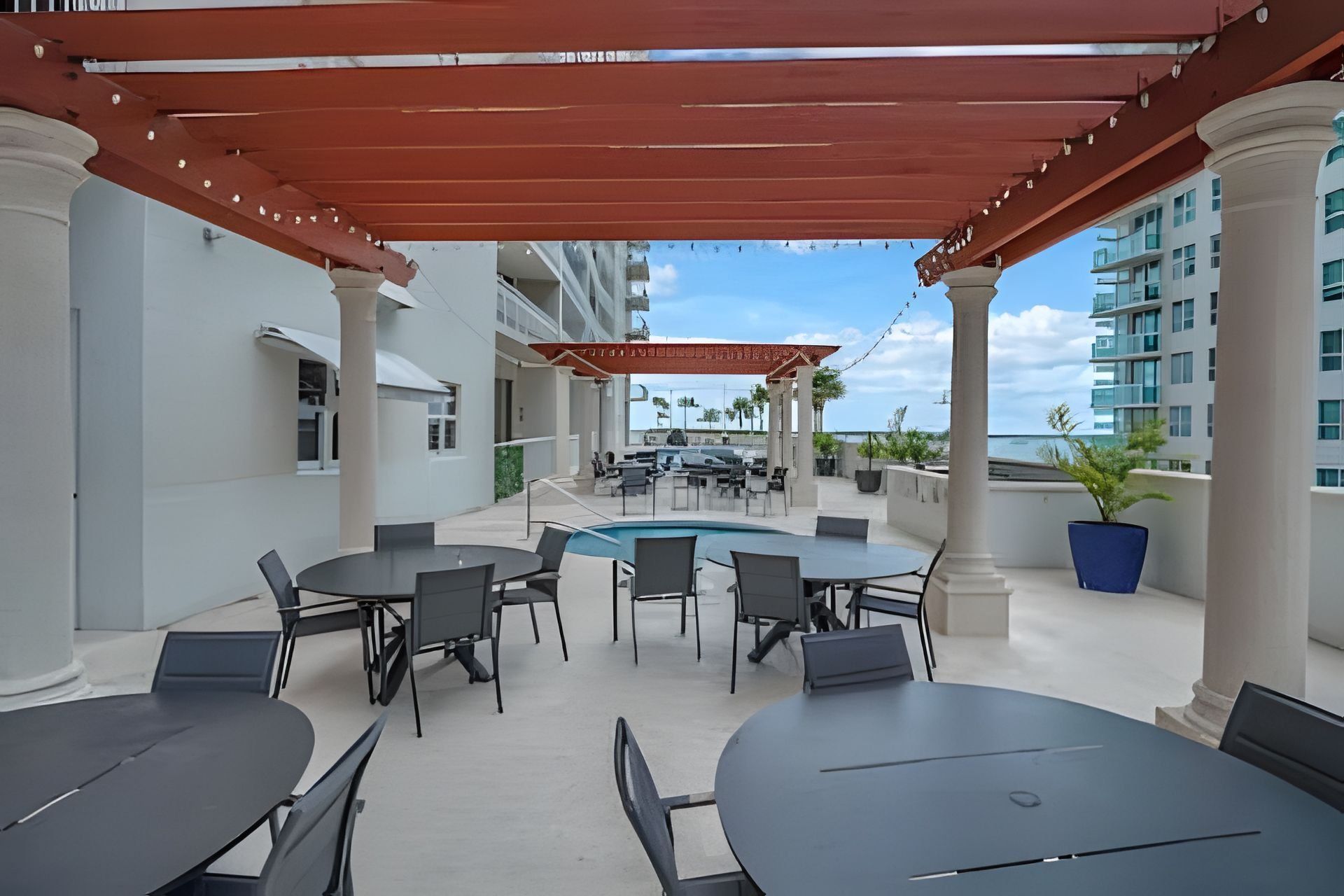 Área de terraço ao ar livre com mesas e cadeiras redondas, piscina e vista para o oceano.