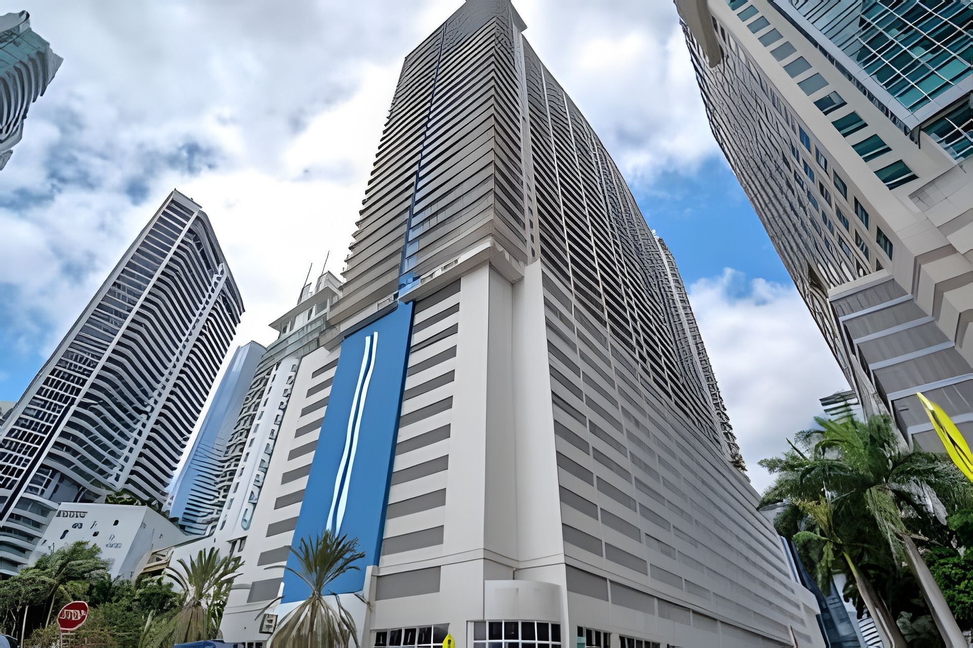 Een modern hoog gebouw met een opvallende blauwe verticale strook, omringd door andere hoge gebouwen en palmbomen.