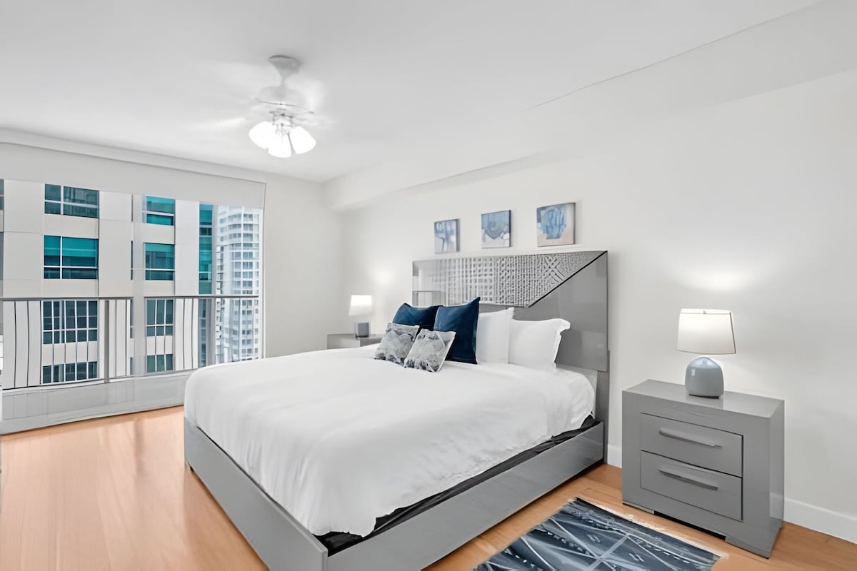 Un dormitorio moderno con una cama grande, mesitas de noche y vistas a edificios altos.