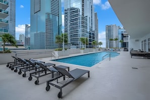 Un área de piscina en la azotea con sillones de descanso y vistas a modernos rascacielos.
